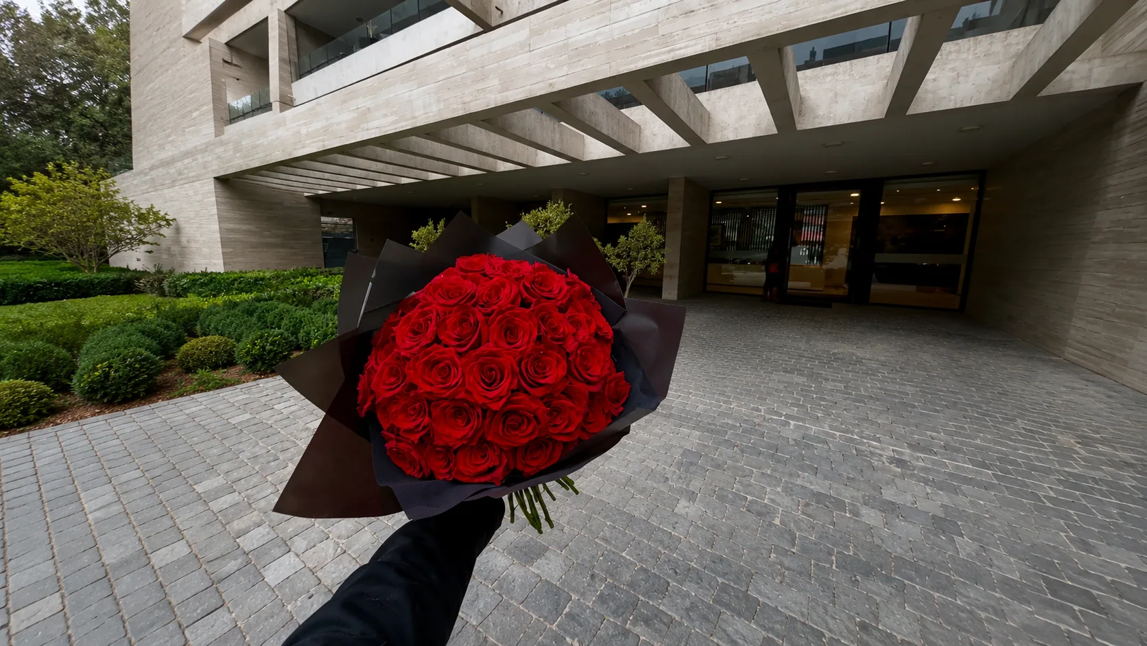 Ramo de rosas rojas en papel negro, sostenido en mano frente a la entrada de un edificio